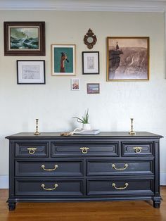 an old dresser is painted black with gold handles and brass pulls in front of pictures on the wall