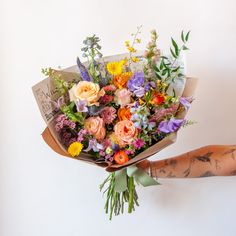 A tattooed arm holds a bouquet wrapped in floral newspaper and brown kraft paper in front of a white backdrop. It's filled with pink, orange, yellow, and purple flowers, including piccolini daisies, roses, veronica, sweet pea, clematis, delphinium, oncidium orchid, and lisianthus. Wildflower Bridal Bouquets, Wildflower Wedding Theme, Wildflower Wedding Bouquet, Boquette Flowers, Spring Wedding Flowers, Colorful Bouquet, Wildflower Bouquet, Beautiful Bouquet Of Flowers, Wildflower Wedding