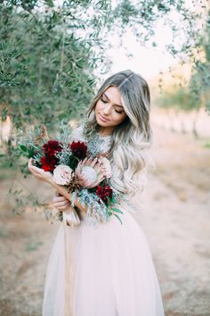 a woman in a white dress holding a bouquet of flowers and greenery with an olive tree behind her