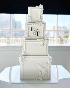 a three tiered white wedding cake with the initials on each layer and flowers at the bottom