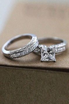 two wedding rings are sitting on top of a box, one has a princess cut diamond