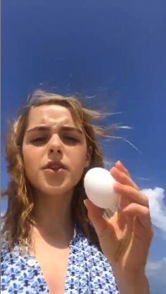 a girl holding an egg in her hand and looking at the camera with blue skies behind her