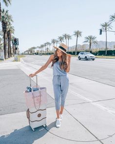 a woman is standing on the sidewalk with her luggage and smiling at the camera while wearing a hat