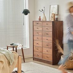 a woman walking through a living room next to a tall wooden dresser with drawers and baskets on it