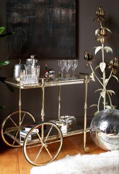 a gold bar cart with wine glasses on it in front of a mirror ball and potted plant