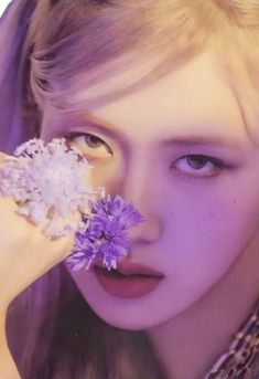 a woman holding a purple flower in her right hand and looking at it through the lens