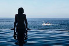 a woman sitting on the edge of a swimming pool looking out at an ocean with a boat in the distance