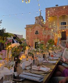 people are sitting at a long table in front of an old building with lights strung from it