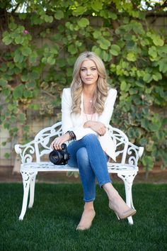 a woman sitting on a white bench in front of a green bush with her camera