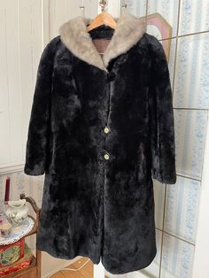 This coat will keep you warm - and looking glamorous! - in any weather. It's made of pure black, thick and plush mouton fur, adorned with a luxuriant soft grey mink fur collar. It fastens with the original decorative buttons and elastics in the front, has two front pockets and one inside pocket, and is fully lined with medium brown silky lining. The measurements, taken with the coat lying flat, are: shoulder to shoulder, 19 inches; armpit to armpit, 23 inches; sleeves, 23 inches; length, 42 inches; bottom edge, 42 inches. There's a bit of wear in the lining of one armhole, but otherwise the coat is in very good condition. Black Long Winter Fur Coat, Black Long Faux Fur Coat, Classic Black Fur Coat With Faux Fur Trim, Formal Black Faux Fur Coat, Classic Black Fur Coat With Faux Fur Lining, Elegant Black Fur Coat With Faux Fur Trim, Mink Color Long Fur Coat With Faux Fur Lining, Black Formal Fur Coat With Faux Fur Trim, Vintage Black Fur Coat With Faux Fur Lining