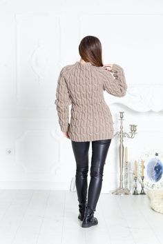 a woman standing in front of a white wall wearing black pants and a beige sweater