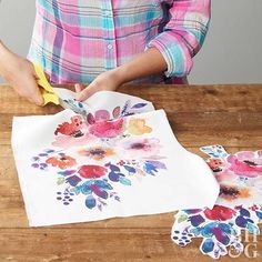 a woman is painting flowers on a piece of paper with scissors and glue while sitting at a wooden table