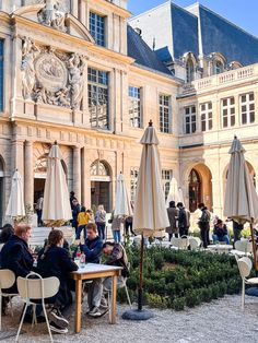 people sitting at tables in front of a large building with many windows and umbrellas