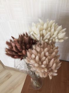 three dried flowers in a glass vase on a wooden floor next to a white wall