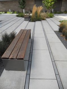 a wooden bench sitting on top of a cement walkway next to grass and bushes in front of a building