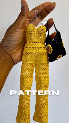 a hand holding a small doll wearing yellow crochet pants and top with a black purse