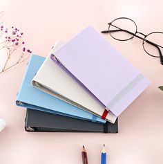 four notebooks with pens and glasses on a pink surface next to flowers, pencils and eyeglasses