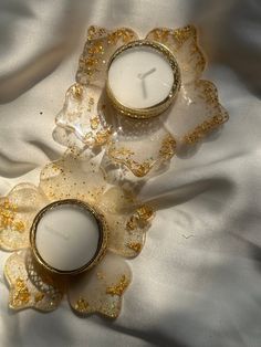 two candles sitting on top of a white cloth next to each other with gold accents