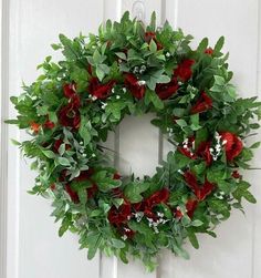 a wreath with red flowers and green leaves hanging on a white front door, surrounded by greenery