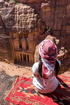 a woman sitting on top of a red rug next to a cliff with a building in the background