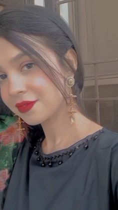 a woman with dark hair wearing red lipstick and gold earrings, standing in front of a window