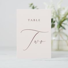 a table card with the word two written in cursive writing on it next to a vase filled with flowers