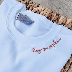 a white t - shirt with the word hey pumpkin embroidered on it sitting in a woven basket