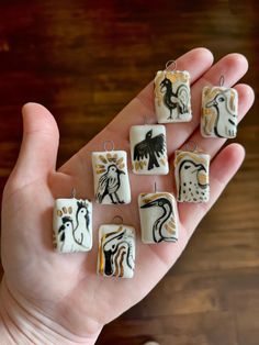 a hand holding nine small square shaped ceramic pendants with different designs and colors on them