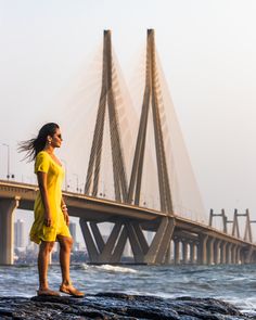 a woman in yellow dress standing on rocks near the water with a bridge in the background