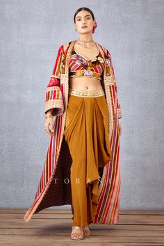 Ochre yellow cape with stripe print and gota patti, mirror embroidered motifs. Comes with floral print bralette and dhoti pant.
Component: 3
Pattern: Print and Embroidery
Type Of Work: Digital Print, Gota Patti, Hand Adda Work and Zari Embroidery
Neckline: Leaf
Sleeve Type: Bralette: Sleeveless and Cape: Flared
Fabric: Silk Velvet and Silk Crepe
Color: Yellow
Other Details: 
Gota patti trim on the bralette hems
Dhoti pant with embroidered waistband
Open front cape
Low back with tie up and tassel Bohemian Style Designer Sets With Zari Work, Multicolor Fitted Dabka Choli, Bohemian Dabka Lehenga For Festive Occasions, Festive Bohemian Lehenga With Dabka Detailing, Festive Bohemian Lehenga With Dabka, Bohemian Dabka Straight Kurta Set, Bohemian Straight Kurta Set With Dabka Detailing, Bohemian Palazzo Set With Dabka Work For Diwali, Bohemian Chanderi Palazzo Set For Festive Occasions