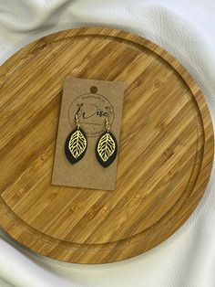 a pair of black and gold leaf earrings sitting on top of a wooden tray next to a white cloth