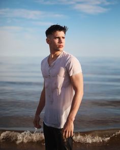 a young man standing on the beach next to the ocean looking at the camera with his hand in his pocket