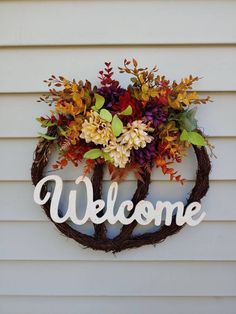 a welcome sign hanging on the side of a white house with autumn foliage and flowers