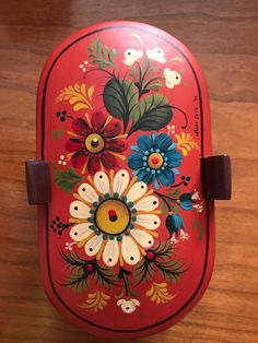 a red box with flowers painted on the front and sides, sitting on a wooden surface