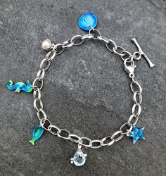 This silver charm bracelet is made from Sterling Silver and Enamel . The Silver Belcher chain which holds various enamel charms that capture the wonders of the ocean has  links that are each approx: 8mm long. It has an adjustable trigger so fits any size wrist. The charms are a starfish, shell, fish, sea horse and a Pearl and a tear-drop, faceted Aquamarine. The measurement is approximately: 8.5 inches but is adjustable. Enamel Bracelet Jewelry With Lobster Clasp, Enamel Bracelet With Lobster Clasp, Nickel-free Enamel Bracelet As A Gift, Enamel Charms Bracelets For Gift, Enamel Charms Bracelet, Silver Enamel Charms With Lobster Clasp, Blue Charm Bracelet With Dangling Charms As Gift, Themed Bracelet With Lobster Clasp, Themed Jewelry With Lobster Clasp