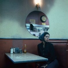 a woman sitting at a table in a restaurant with a mirror above her head and a clock on the wall