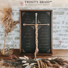 a wooden cross sitting on top of a table next to dried flowers and feathers in front of a sign that reads trinity braid