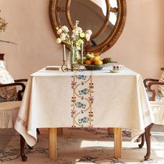 a table with flowers and fruit on it in front of a round mirror, near two chairs