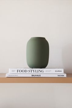 a green vase sitting on top of a book next to a white wall and wooden shelf