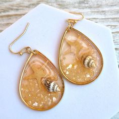 "These exquisite Boho Handmade Coral Peach Translucent Resin Seashell Starfish Ocean Themed Sealife Teardrop Dangle Earrings are perfect for adding a touch of seaside charm to your jewelry collection. Each earring features a beautifully crafted translucent resin teardrop in a refreshing mint color, reminiscent of the ocean's tranquil hues. Encapsulated within the resin are delicate seashells and a whimsical starfish, capturing the essence of the beach and its mesmerizing sealife. Measuring at a comfortable 1.75\" (4.45 cm) drop, these earrings offer an elegant and eye-catching length, allowing them to sway gently with your movements. The teardrop shape adds a graceful touch, enhancing their overall appeal. These earrings also make fantastic vacation gifts, serving as a delightful reminder Gold Dangle Teardrop Earrings For Beach, Gold Teardrop Earrings For Summer, Beach Dangle Teardrop Earrings With Ear Wire, Summer Beach Teardrop Earrings, Gold Starfish Shell For Beach, Handmade Teardrop Earrings For Beach, Black Gold Necklace, Boho Handmade, Teardrop Dangle Earrings