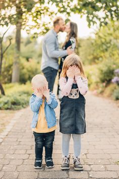 two children covering their eyes as they stand on a brick path in front of trees