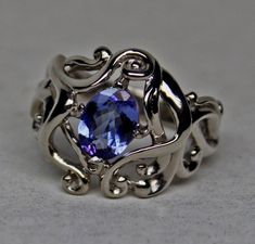 This ring was inspired by the Art Nouveau movement as well as Celtic interlace designs. This ring was designed and made by George Postgate. It was hand sculpted in wax, and then carved in metal, after it was cast in 14KPA white gold. The ring is set with a beautiful, natural, oval faceted; tanzanite that weighs 1.01 cts. The stone is an very fine example that is flawless to the eye. The stone is a nice medium blue color with a nice dance of secondary colors that appear as the stone is moved. Tan Unique Sterling Silver Sapphire Ring For Formal Occasions, Unique Formal Sapphire Ring In Sterling Silver, Elegant Hand Forged Sapphire Ring Gift, Elegant Hand Forged Sapphire Ring For Anniversary, Unique White Gold Sapphire Ring, Unique White Gold Sapphire Ring For Formal Occasions, Art Nouveau White Gold Rings For Formal Occasions, Victorian Gothic Jewelry, White Gold Set