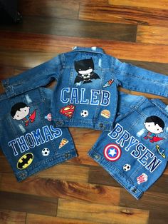 three children's jean jackets with superman and captain america patches on them, sitting on a wooden floor