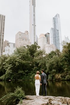 a bride and groom standing on rocks in the middle of a lake surrounded by tall buildings