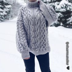 a woman standing in the snow wearing a gray sweater and black pants with her hands on her head