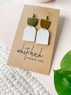 the white and gold square earrings are on top of a card next to a plant