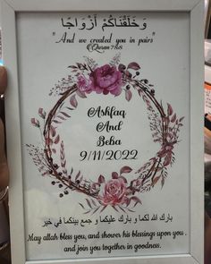 a hand holding up a white frame with pink flowers and leaves in arabic writing on it