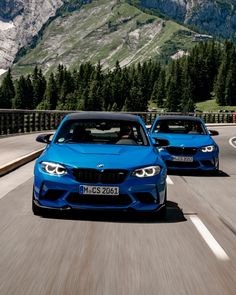 two blue bmw cars driving down the road in front of some mountains and pine trees