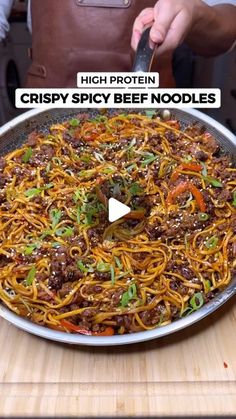 a person is cutting up noodles in a skillet on a wooden table with the words, high protein crispy spicy beef noodles