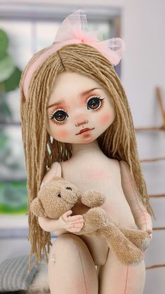 the doll is holding a teddy bear in her hands and wearing a pink bow on her head